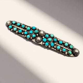 A silver-toned Southwestern-style turquoise belt buckle or brooch, featuring multiple round turquoise stones and sunburst details, photographed on a light beige background with soft diagonal shadows.