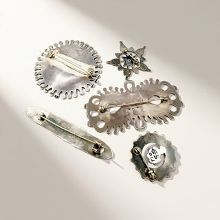 Five silver-toned vintage brooches and pin backs arranged on a warm off-white background with soft diagonal shadows. The pieces include scalloped circular, floral, and elongated openwork designs, with one pin back showing a silver clasp and another showing the interior pin mechanism.