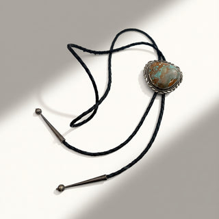 A black cord bolo tie necklace featuring a large oval turquoise stone in a silver-toned bezel setting, displayed on a white surface with soft diagonal shadows.