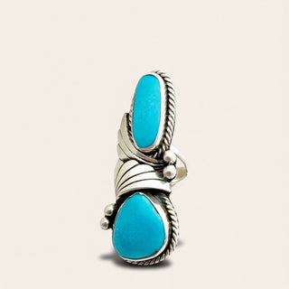 A silver ring featuring two large oval turquoise stones set in a stacked, wrapped design with textured rope borders and small bead accents, shown on a light beige background.