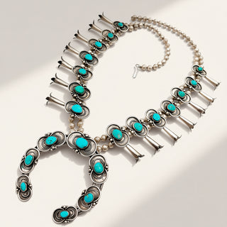 A silver-toned Southwestern-style necklace featuring multiple turquoise cabochon stones, small white pearl beads, and elongated silver bell-shaped drops, arranged on a light beige background with soft shadows.