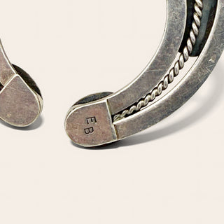 Close-up of a silver-toned curved jewelry with a twisted rope detail and engraved initials 'EB' on the clasp, shown on a light beige background.