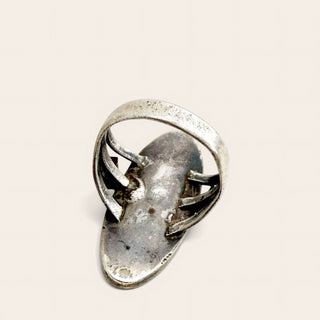 A silver ring with a wide, textured band and three curved, claw-like side elements holding a central oval stone, shown on a plain light beige background.