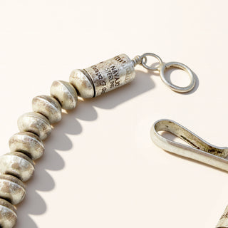 A close-up of a silver-toned jewelry clasp component featuring a cylindrical engraved bead with text and a round ring, alongside part of a metal loop on a light beige background.