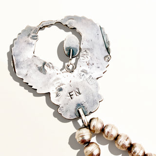 A close-up of a silver-toned metal pendant or charm with a scalloped irregular edge, stamped initials 'FN', and a dangling beaded strand in metallic gold and silver tones on a plain light background.