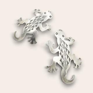 Two silver gecko-shaped earrings with intricate cutout patterns, displayed on a light beige background.