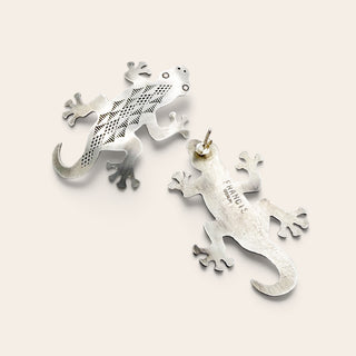 Two silver gecko earrings on a light beige background. One earring features a textured, perforated pattern, while the other shows the polished back.