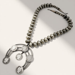 A silver necklace featuring a strand of polished and textured gray beads with a large circular pendant adorned with five coin-like medallions showing profile portraits, photographed on a light neutral background with soft shadows.