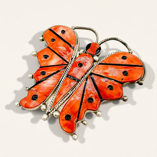 A butterfly-shaped brooch featuring vibrant orange inlay panels with black accents and silver-toned metal borders, photographed on a light beige background.