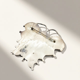 A silver-toned abstract leaf-shaped brooch with a pin fastener, displayed on a light beige background with soft diagonal shadows.
