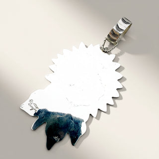 A sterling silver pendant featuring a stylized raven silhouette with a textured, sunburst-like metallic top edge and a dark blue inlay at the bottom. The pendant is suspended from a matching silver bail decorated with black engraved eye motifs, set against a neutral light gray background.