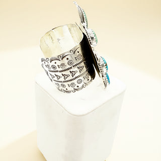 Decorative silver cuff with intricate patterns on a white stand against a beige background