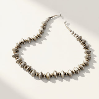 A beaded necklace made of small metallic disc-like beads with a polished silver-tone finish, arranged in a curved shape on a light cream background with soft shadows.