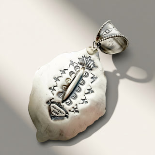 A silver pendant with intricate hand-stamped geometric and tribal patterns, featuring a central elongated stone and a decorative bail, shown on a neutral light gray background with soft shadows.