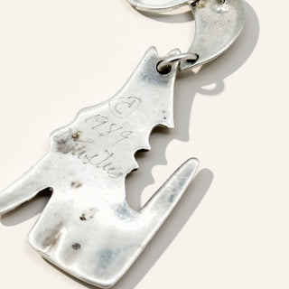 A close-up of a silver charm with engraved markings, shown on a light cream background.