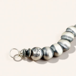 A close-up of a metallic beaded necklace featuring brushed silver and dark gray round beads, with one bead engraved 'MK' and 'STAINLESS' on a light beige background.