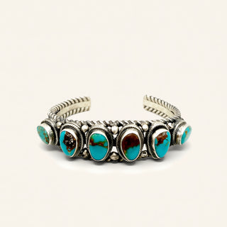A sterling silver cuff bracelet featuring six oval turquoise stones with brown matrix, set in a textured bezel setting on a light cream background.