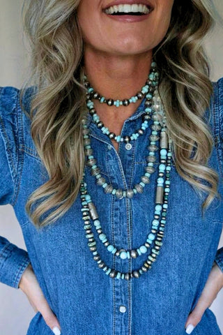 Woman wearing a blue denim shirt and multiple beaded necklaces with a neutral background