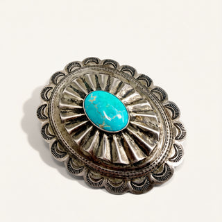 A sterling silver oval pin featuring a central turquoise cabochon stone, set against a plain off-white background.