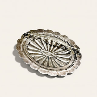 An oval silver-toned metal brooch with a scalloped edge and a sunburst radial pattern, photographed on a plain light beige background.