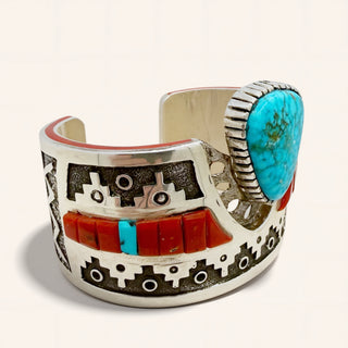 A wide sterling silver cuff bracelet featuring geometric black and white inlay patterns, a row of red coral inlays, and a large oval turquoise stone on the side.