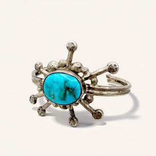A sterling silver cuff ring featuring a central oval turquoise cabochon surrounded by decorative, spiky silver details, set against a plain off-white background.