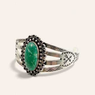 A sterling silver cuff bracelet with a large oval green stone centerpiece, surrounded by a scalloped beaded border, on a light cream background.