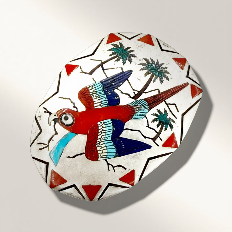 A hand-painted ceramic plaque featuring a red and blue parrot, green palm trees, and red triangles on a white background with black geometric borders.