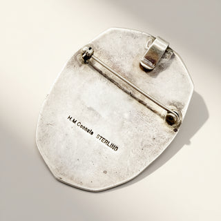 A vintage sterling silver pin or brooch with a flat, shield-like shape and a visible metal clasp mechanism on the reverse side. The surface shows engraved text reading 'H.