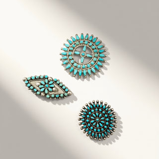 Three turquoise and silver Native American style jewelry pieces, including two circular sunburst designs and one teardrop-shaped piece, arranged on a light beige background with soft shadows.