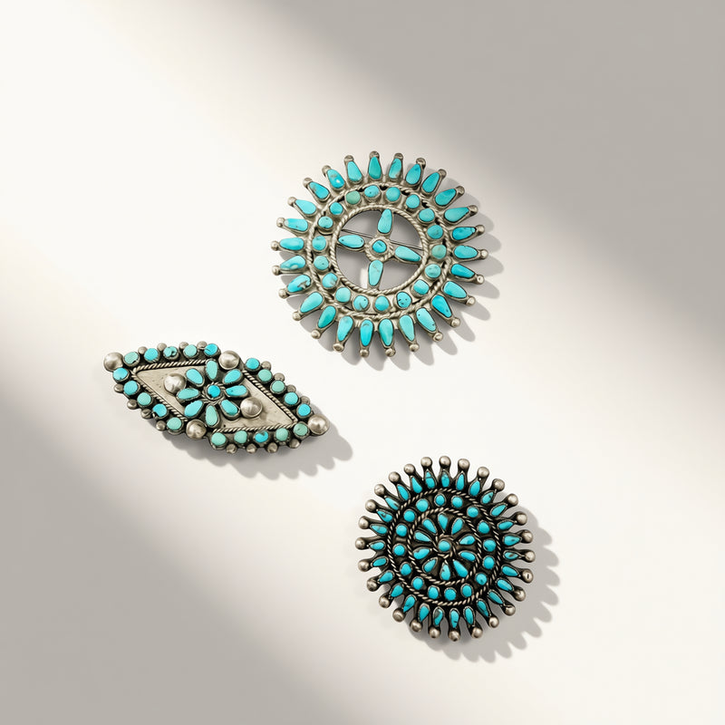 Three turquoise and silver Native American style jewelry pieces, including two circular sunburst designs and one teardrop-shaped piece, arranged on a light beige background with soft shadows.