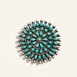 A circular silver-toned jewelry piece featuring a geometric pattern of small turquoise stones and metallic bead-like accents, photographed on a plain light background.