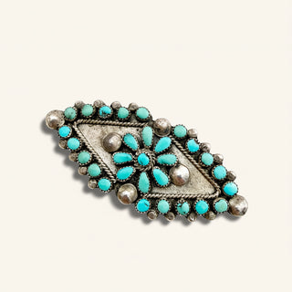 A vintage-style silver-toned belt buckle featuring a floral arrangement of turquoise stones and silver beads on a cream-colored background.