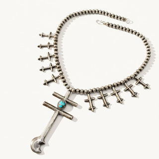 A silver-toned Southwestern-style necklace featuring a beaded chain, multiple small cross charms, and a larger central cross pendant with a turquoise stone, displayed on a clean off-white background.