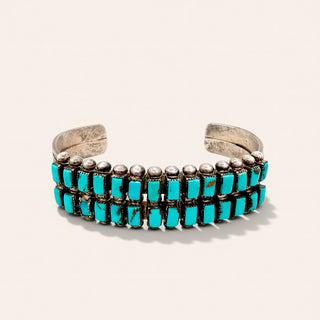 A silver cuff bracelet featuring three rows of rectangular turquoise stones and small round silver beads, set against a light beige background.
