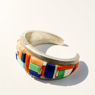 A sterling silver cuff bracelet featuring a colorful inlay of geometric mosaic tiles in orange, blue, green, and white, photographed on a clean white background with a soft shadow.