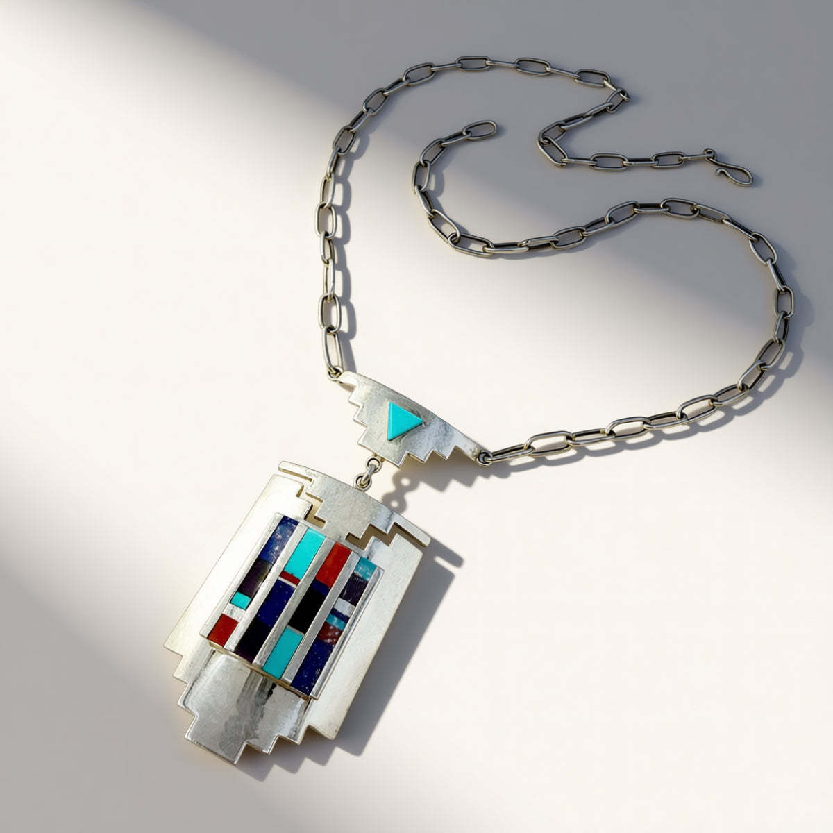 A silver-toned geometric pendant necklace with a multi-colored inlay design featuring turquoise, red, blue, and white accents, displayed on a clean white background with soft shadows.
