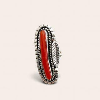 A Southwestern-style sterling silver ring featuring a large, polished red coral cabochon in an elongated oval setting, adorned with a feather motif and beaded borders on a light beige background.