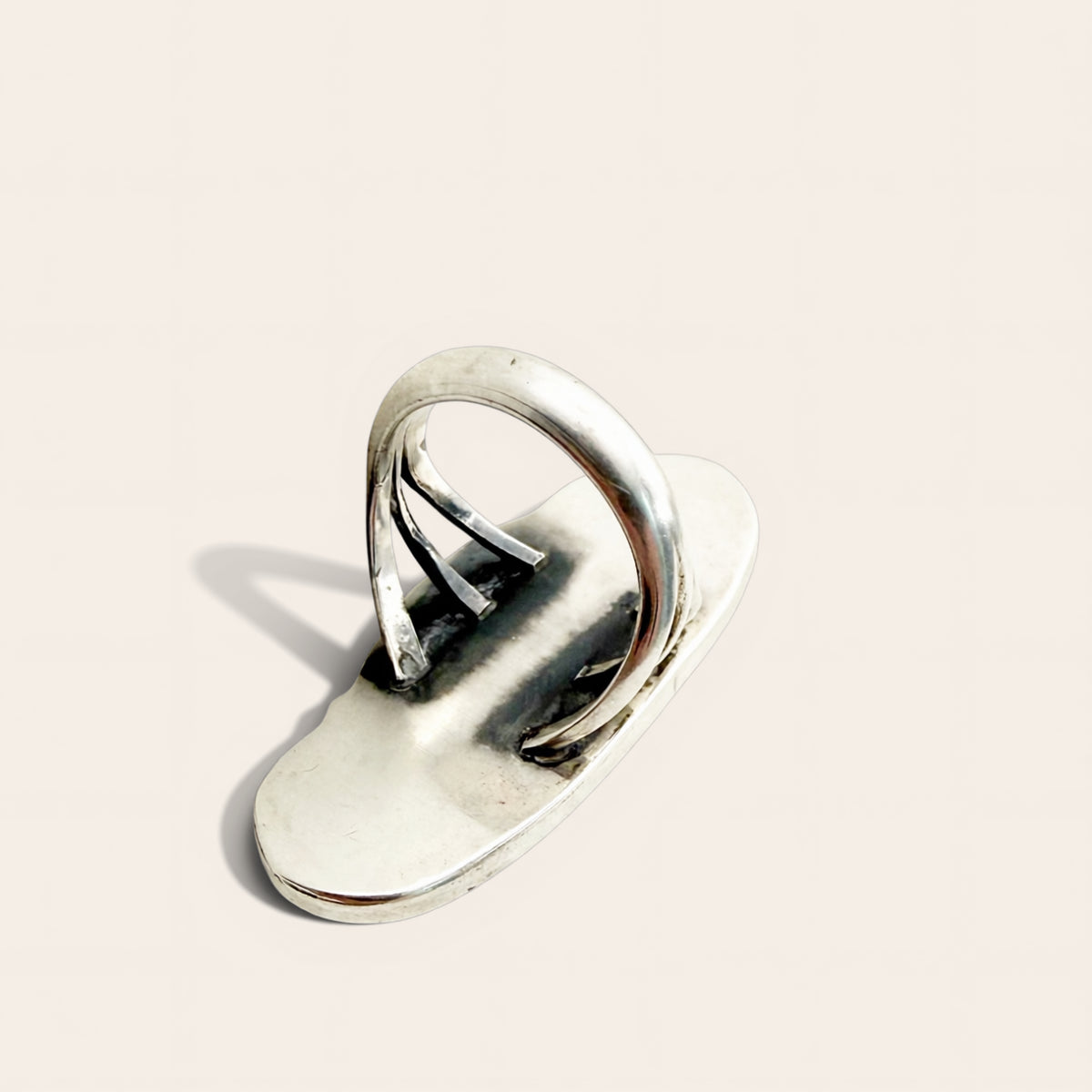 A polished silver-toned metal ring with a flat, elongated oval base and three parallel looped bands rising from the center, photographed on a clean warm off-white background.