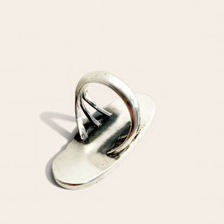 A polished silver-toned metal ring with a flat, elongated oval base and three parallel looped bands rising from the center, photographed on a clean warm off-white background.