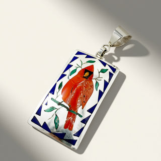 A rectangular silver pendant featuring an inlay design of a red cardinal bird on a branch, surrounded by blue and white geometric patterns, shown on a light background.