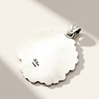 A sterling silver pendant with a scalloped, organic wavy edge and a small bail, shown on a warm beige background with soft diagonal shadows. The inside of the pendant is stamped with 'sterling'.