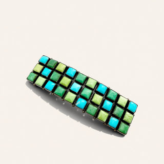 A rectangular woven hair barrette featuring a checkered pattern of turquoise blue and lime green resin tiles set in black metal, shown on a light beige background.