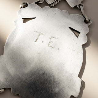 Close-up of a silver pendant with a scalloped border and cutout details, featuring the engraved initials 'T.E.