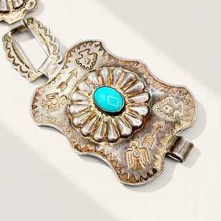 A close-up of a silver-toned Southwestern-style pendant or bracelet piece with a turquoise cabochon in the center, photographed on a light neutral background with soft shadows.