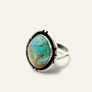 A sterling silver ring featuring a large, oval-shaped turquoise stone with natural brown matrix veining, set in a decorative bezel with black accents on a light cream background.