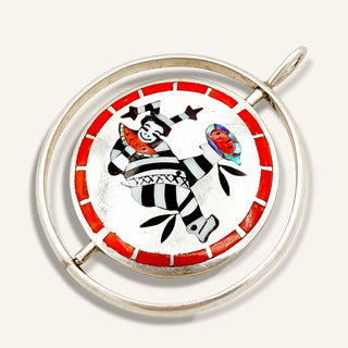 A circular silver-toned pendant featuring a hand-painted Inuit-style design with a black-and-white striped figure, a red border, and small colorful details, shown on a light beige background.