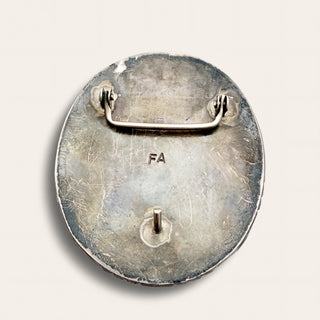 The back of a round silver-toned pin badge with a metal clip and the letters 'FA' stamped in the center, shown on a light beige background.