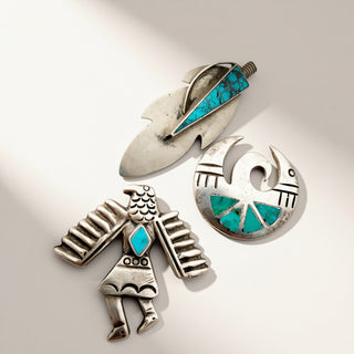 Three silver Native American style jewelry pieces with turquoise inlay, including a bird figure, a circular bird emblem, and a feather-shaped pendant, displayed on a light beige background.