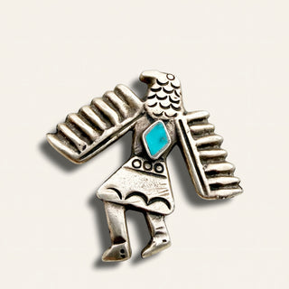 A silver-toned Southwestern-style eagle and human figure brooch with a turquoise inlay, shown on a light beige background.